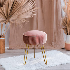 Pink velvet stool with gold legs in a room with decorative plants and a beige curtain.