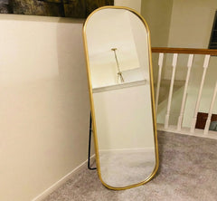 Gold-framed standing mirror leaning against a wall in a room with a staircase.
