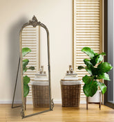 Decorative standing mirror with ornate design in a room with plants and wicker baskets.