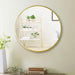 Round gold-framed mirror on a wall with a shelf and plants reflected in it.