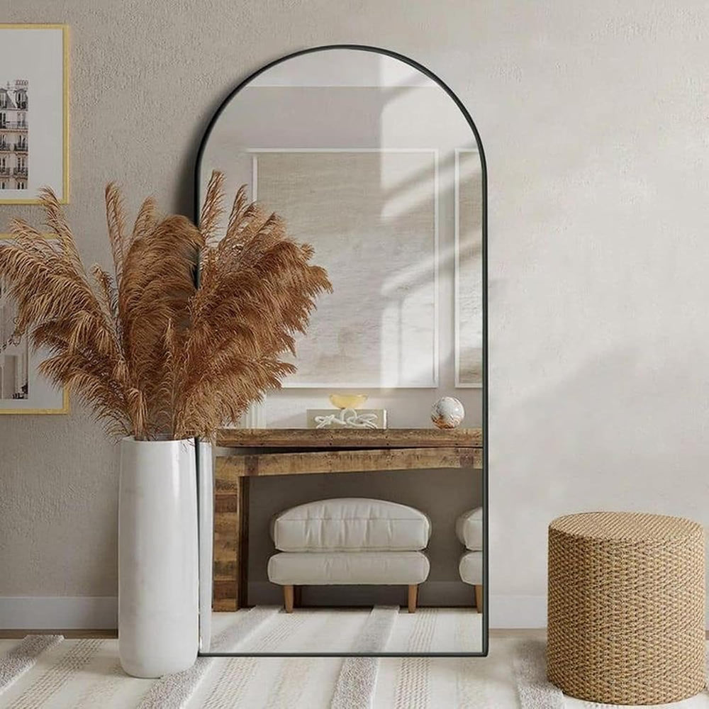 Large arch-shaped mirror in a room with a vase of pampas grass, a wooden console table, and a wicker stool.