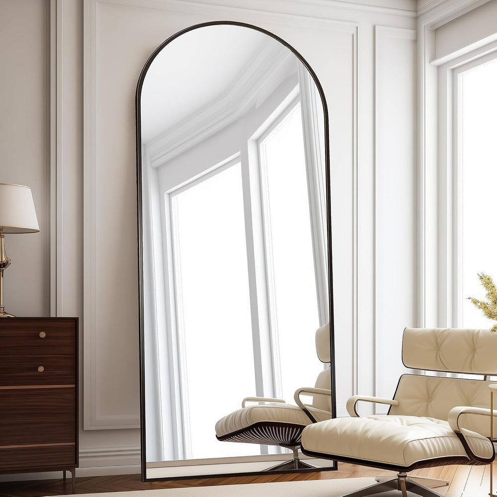 A tall floor mirror with a black arched frame stands in a bright, modern room with white paneled walls. The mirror reflects large windows, and a cream tufted lounge chair is partially visible in the reflection.