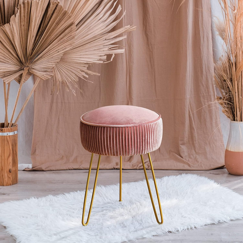 Pink velvet stool with gold legs in a room with decorative plants and a beige curtain.