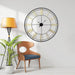 Large wall clock on a white wall with a wooden chair and plant in the foreground.