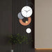 Modern wall clock with decorative elements on a wall with a plant in the foreground.