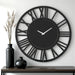 Large black wall clock with Roman numeral design on a white wall, accompanied by two black vases.