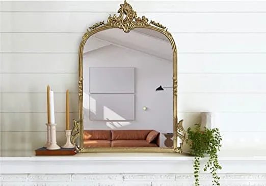 Decorative gold mirror on a white wall with candles and a plant on a shelf below.